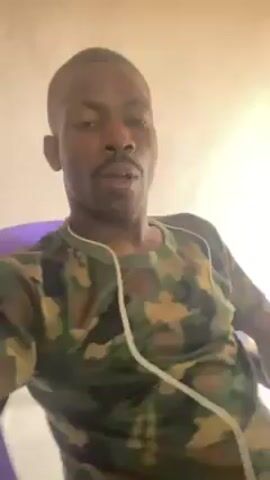 The video shows a man from the chest up sitting in a purple chair. He is wearing a camouflage t-shirt and has white headphones plugged in. He gestures with his right hand, flexing his bicep at one point, and then runs his hand over his shaved head. He has a mustache and appears to be looking directly at the camera. The background is a plain white wall.