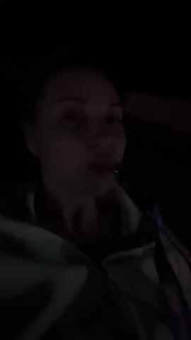 A person, likely a woman, is smoking a hookah in the dark.  She is wearing what appears to be a light-colored jacket. The hookah hose is dark, and the smoke exhaled is light gray. The environment is very dark, with minimal light revealing her face and upper body. The background is completely blacked out.