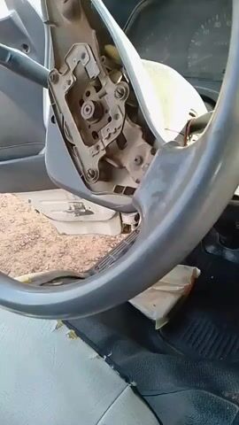 The video shows the interior of a vehicle, focusing on the steering column area. The steering wheel appears to be partially disassembled or removed, revealing the mechanical components beneath. A grey plastic shroud is lifted, exposing metal brackets, screws, and wiring. The steering wheel itself, a large grey circular object, is prominently featured in the foreground. To the left, a turn signal lever is visible. The background shows parts of the dashboard, including a speedometer, and the vehicle's interior, such as a seat and floor mat. The lighting suggests it is daytime, and the environment outside the vehicle appears to be a dry, outdoor area with dirt or gravel visible through an open door. There are no people visible in the video.