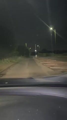 The video shows a first-person perspective from inside a car driving at night. The dashboard of the car is visible at the bottom of the frame, with dust on the windshield. The road ahead is illuminated by the car's headlights and several streetlights lining the road. On the left, there are trees and bushes, and a small road sign is partially visible. On the right, there are red and white diagonal road markings, possibly indicating a pedestrian crossing or a traffic calming measure, and a yellow sign. The sky is dark, indicating nighttime. The road appears to be a two-lane road with a broken white line in the center, and solid white lines on the edges. The overall scene suggests driving through a suburban or semi-rural area at night. No people are visible, and no discernible text is present.
