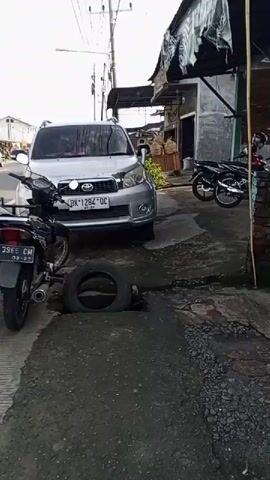 A silver SUV, license plate BK 1284 OC, is parked on a rough, cracked concrete surface. In the foreground, a detached tire lies partially within a square opening in the ground, possibly a drain or manhole. To the right of the SUV, two motorcycles are parked. One is black and red, and the other is black. Beyond the motorcycles, a building with a corrugated metal roof is visible. The overall environment appears to be an outdoor, possibly residential or commercial, area with unkempt surroundings.