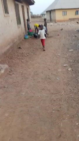 The video shows an outdoor scene in what appears to be a rural area. The ground is dirt. Several children are present, and one is walking toward the camera wearing a white shirt and red pants. Further back, three individuals are gathered around two large black basins. They appear to be washing or rinsing something. There are several containers, including a green basket and a blue basket, and various dishes scattered around the area. There are buildings visible in the background, with one close to the camera and two more in the distance.