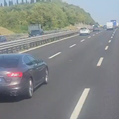 The video shows a gray Maserati Ghibli driving on a highway. The license plate is GT-702-LX. The car is in the right lane. There are other cars and trucks on the highway, and a hill with trees on the left. The sky is blue and it appears to be daytime.