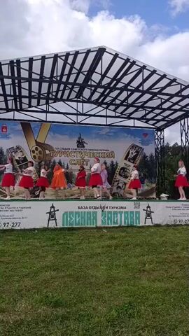 A group of seven young females are performing a dance routine on a raised outdoor stage under a cloudy sky. The dancers are dressed in matching red and white outfits with white skirts and red tops, except for one dancer in the center who is wearing an orange dress. They are positioned across the stage, performing synchronized movements. Behind them, a large banner displays the text "ПО СЛЕДУ СОВЕТСКОГО КИНЕМАТОГРАФА ТУРИСТИЧЕСКИЙ СЛЕТ" (Following the Soviet cinematography tourist gathering) along with images of film reels and trees. A white banner with green lettering reading "ЛЕСНАЯ ЗАСТАВА" (Forest Outpost) is visible at the bottom of the stage. The stage is constructed with a metal frame and a translucent roof. The area in front of the stage is covered in green grass.