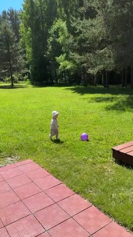 A baby in a white shirt, striped shorts, and a white sun hat stands in a green grassy field on a sunny day. The baby picks up a small purple ball and holds it for a moment, then looks around. Trees are visible in the background, and a red tiled patio is in the foreground.