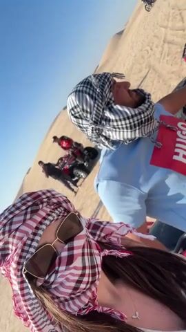 A low-angle shot shows a person wearing sunglasses and a red and white patterned headscarf looking at the camera. Behind them is another person wearing a blue shirt with the word "HUGO" in red and a black and white checkered headscarf. The background is a desert landscape with several red ATVs lined up on the sand dunes under a clear blue sky. Some other people are seen standing near the ATVs in the background.