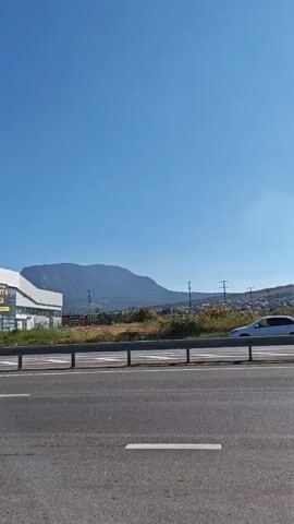 A white car drives on a highway next to a metal guardrail. In the background, there are houses and hills under a clear blue sky with the sun shining. The highway has white lane markings.