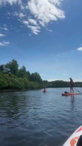 The video shows a wide, calm body of water, likely a lake or river, on a sunny day with a blue sky and scattered white clouds. Dense green trees line the far bank of the water. In the foreground, the edge of a white and orange paddleboard is visible. Further out on the water, two people are paddleboarding. One person, on the right side of the frame, is standing on a red paddleboard, wearing a dark top and shorts, and holding a paddle. Another person, on the left side of the frame and further away, is sitting on a dark paddleboard and paddling with a long oar. The person on the left appears to be moving their arms in a way that suggests they are actively paddling or adjusting their position on the board. The overall scene depicts a recreational activity on a natural body of water.