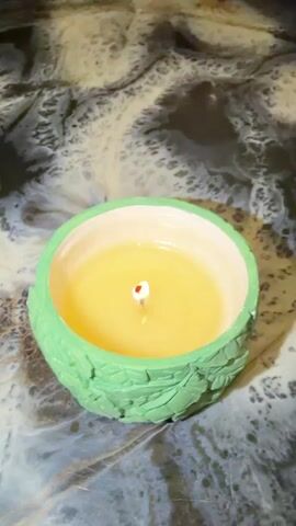 A lit candle in a green decorative holder is shown. The candle wax is yellow. The holder is decorated with leaves. A hand places a white lid with a yellow center and white petals on top of the candle holder, covering the candle. The background is a dark, swirled surface.