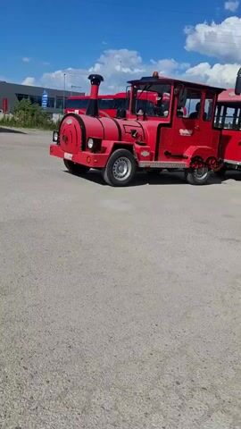 The video shows a bright red trackless train, with a bus parked in the background. The "train" is designed to resemble a steam locomotive, with a smokestack and rounded front that imitates a boiler. The train has a passenger car attached to it. The vehicle appears to be parked on a paved lot on a sunny day. There are a few white clouds in the blue sky.  The location looks like it could be a bus or train depot.