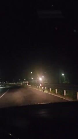 The video, filmed from inside a vehicle at night, shows a dark road ahead illuminated by the vehicle's headlights. A white line is visible on the left side of the road. To the right, a metal guardrail runs parallel to the road. Beyond the guardrail, a long structure with multiple illuminated windows, resembling train cars or a building, is visible. Scattered bright lights, possibly streetlights or distant structures, are visible in the background. Briefly, a distinct, bright, cross-shaped light appears high in the dark sky before fading from view. The top of the vehicle's dashboard and the bottom of the windshield frame are visible in the foreground.