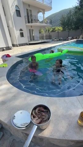 A person's hand holds up a raw shrimp as three young girls play in a swimming pool. Two girls are in the water near the edge, and one is on a bright green inflatable float in the background. A pot filled with raw shrimp sits on the edge of the pool in the foreground. The setting appears to be a sunny day in a backyard with a house and trees visible.