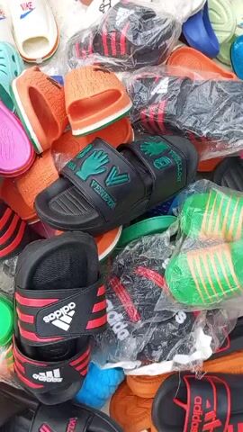 The video shows a pile of various footwear, including slides and clogs. There are pairs of pink clogs, Adidas slides in multiple color combinations such as black with red stripes, and green and orange striped slides in plastic packaging. There is a black pair of slides with the word "Veneta" and green hand-shaped designs. A loud, drilling noise is audible.