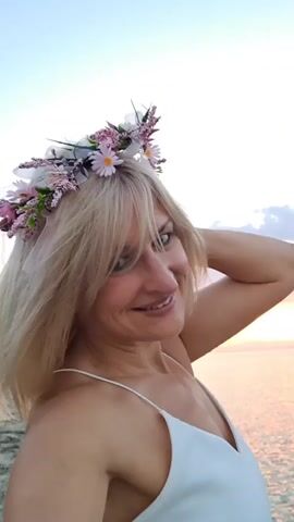 A close-up, vertical video shows a woman with blonde hair and a flower crown on her head. She is wearing a white dress with spaghetti straps and smiling at the camera. She is holding her right hand to the back of her head. The background is a blurry body of water with a sunset.