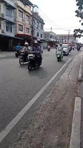 A street scene with buildings on the left and a sidewalk on the right. Several motorcycles and a silver car are traveling on the paved road. One motorcycle in the foreground has two passengers, a rider and a passenger. Another motorcycle further back has a single rider. The silver car is in the middle of the road. The buildings appear to be commercial with balconies and multiple stories. The sky is overcast.