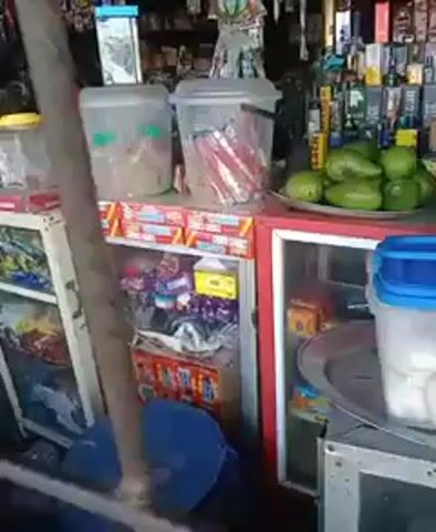 The camera pans across the front of a small store, behind a barred metal fence. The store sells a variety of items, including drinks, snacks, and fruit. There is a refrigerator, a tray of green fruit, and multiple clear containers filled with goods. There is also a selection of alcohol bottles displayed on the top shelf. An indiscernible person is heard speaking in the background.