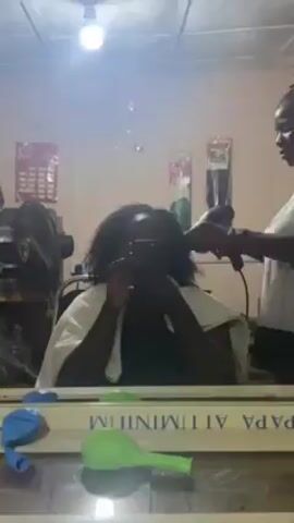 A woman is having her hair dried with a hair dryer by another woman in a salon. The woman getting her hair dried is sitting in a chair and holding up a phone, presumably taking a selfie. She is wearing a dark shirt and a white towel around her shoulders. The woman drying her hair is wearing a white t-shirt and has her hair up in dreadlocks. The salon has a large mirror with decorations around it. A fan, packages of hair extensions, and blue and green balloons are also visible in the room.