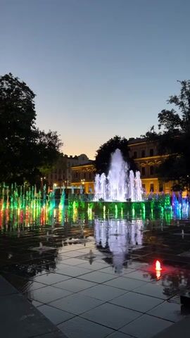 A vertical, low-angle shot shows a fountain display at dusk. The fountain is comprised of numerous jets of water, many of which are illuminated with colorful lights, including blue, green, pink, and white. The water jets are arranged in rows and columns, creating a mesmerizing pattern. In the background, a building with illuminated windows is visible, flanked by trees. The sky is a gradient of deep blue and pale orange, indicating the transition from day to night. The wet surface of the fountain plaza reflects the vibrant lights of the water jets, adding to the magical atmosphere.