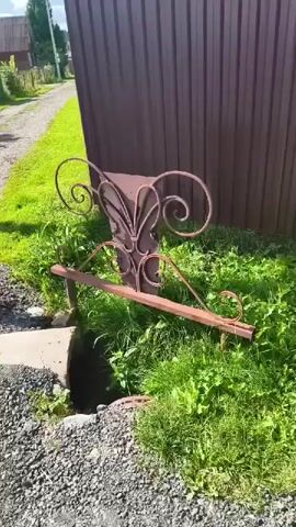 The video shows an outdoor scene with a rusty, ornate metal structure that resembles a bench or planter, situated in front of a tall, brown corrugated metal fence. Green grass and weeds surround the metal structure and grow around a concrete block to its left. The ground is covered in gravel and some dirt. The sunlight suggests it is daytime.