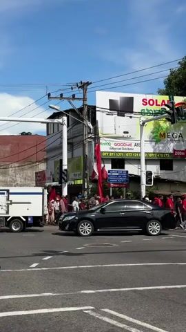 The video captures a lively street scene in what appears to be Delkanda, Sri Lanka, during the daytime under a clear blue sky. A large group of people, many dressed in red clothing and carrying red flags, are actively participating in a procession or protest. They are seen walking across and along the intersection, suggesting a organized public gathering.

Vehicles such as a black car, a blue car, a light blue van, and a motorcycle are present on the road, navigating the flow of people or waiting at the traffic lights. The street features typical urban infrastructure, including traffic lights, power lines, and various commercial buildings.

Prominently visible are large billboards, one advertising "SOLAR MEGA" with a green leaf motif, and another with "GOODHE AFO" along with several phone numbers (077 371 1371, 077 121 8218, 0777 473 273). A sign indicating "200m" with a directional arrow is also visible. In the background, a distinct white stupa-like structure atop a yellow building, adorned with colorful flags, suggests a religious site. Other visible text includes "MANJARI" and various signs in Sinhala script. The scene depicts a dynamic moment of public movement within a busy urban environment.