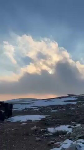 The video shows a landscape with light snow cover under a sky with clouds illuminated by the setting sun. In the foreground, there is a damaged, dark-colored vehicle. In the distance, power lines are visible across the land.