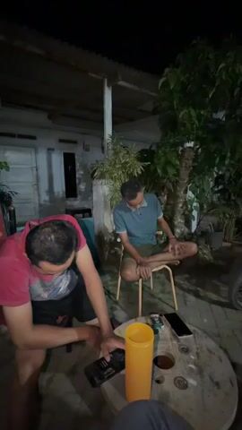 Two men are seated outdoors at night. The man on the left is wearing a red shirt and black shorts. He is leaning forward, holding a black object, possibly a phone, in his left hand. In front of him, on a round table, are a yellow container, a phone, and possibly a pack of cigarettes. The man on the right is wearing a blue shirt and shorts. He is sitting on a stool with his legs crossed and is looking down. The environment appears to be a backyard with a building in the background and trees.