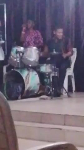 A slightly blurry video depicts a band performing onstage. The scene is dimly lit, making details challenging to discern. 

A drummer is positioned on the right side of the frame, playing a drum set. To the left of the drummer, a person stands singing into a microphone. 

The stage is slightly elevated, with a few steps leading up to it. The background is a dark curtain.

The video quality is poor, with noticeable blurriness and limited visibility due to low lighting.