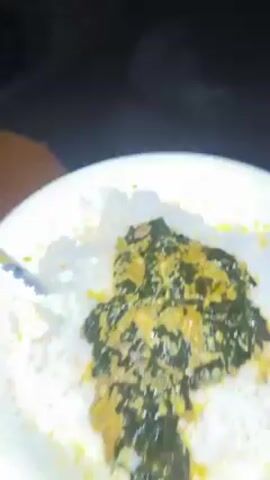 A close-up shot displays a white plate filled with white rice, topped with a portion of green and yellow stew. The camera angle focuses downwards onto the plate. The dish appears to be freshly prepared and is set against a dark background.
