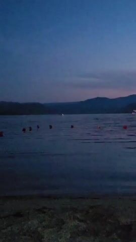 The media file captures a tranquil waterscape at dusk. The scene depicts a calm body of water, likely a lake or reservoir, with gentle ripples reflecting the fading light. Several orange buoys are visible floating on the surface, marking an area of the water. In the distance, a chain of hills or mountains forms a silhouette against the twilight sky. On the far shore, lights from buildings and possibly a boat twinkle, indicating human presence and development. The sky itself is a gradient of deep blue, fading to lighter hues with hints of pink and purple near the horizon, suggesting the transition from day to night. The overall atmosphere is peaceful and serene, evoking a sense of quietude and natural beauty. The foreground shows a sandy or pebbly shore, further grounding the scene on land. The time of day is clearly evening, as indicated by the low light conditions and the colors in the sky. There are no visible people or explicit text in the media file.