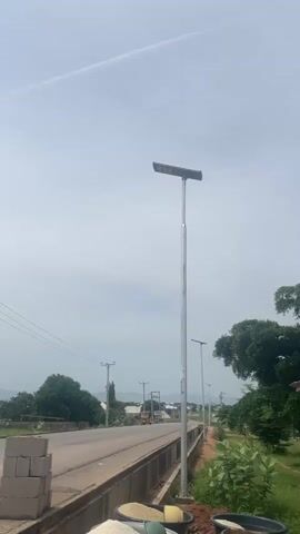 This video captures a serene, daytime scene in Howayi, Nigeria. The focus is on a road with a concrete divider, lined by a solar-powered streetlamp. To the left of the road, a stack of grey concrete blocks sits near the edge, with a glimpse of a yellow truck in the distance. Power lines crisscross the sky, supported by utility poles.

The background reveals a gently rolling landscape with scattered houses, their roofs a mix of white and other muted colors. A prominent palm tree, with its distinctive fan-shaped fronds, stands tall in the middle ground, a symbol of the tropical setting. In the far distance, hazy blue mountains form a majestic backdrop. The sky is overcast, with a faint contrail streaking across it, suggesting it's a moderately cloudy day, likely late morning or early afternoon given the brightness. The overall mood is peaceful and rural, with a sense of development indicated by the new streetlights. No people are visible in the footage.