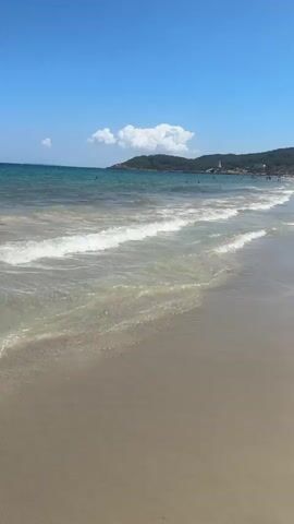 A beach scene on a clear, sunny day. Gentle waves lap onto a sandy shore. In the distance, a hilly coastline is visible, dotted with buildings and a tower. Several people are seen swimming or walking along the beach. The sky is a vibrant blue with a few scattered white clouds. The sand is light brown and appears wet near the waterline, with dry sand further up the beach.