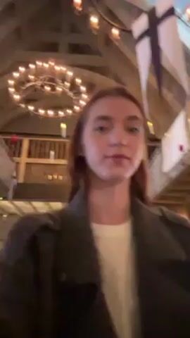 A young woman with long auburn hair is indoors, standing and drinking from a large glass mug. She is wearing a gray wool-like overcoat and a white shirt underneath. She takes a large drink and foam is visible on her upper lip. The location has a large timber frame, and there is a chandelier, flags, and small balconies visible.