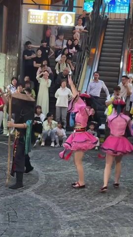 A group of people are gathered to watch a street performance. Two young women in matching bright pink outfits with short skirts and long sleeves are performing a dance. They wear headpieces and face coverings. A man in a black costume with a staff stands next to them. Behind the performers, many people are watching from an elevated area near an escalator. Some people are sitting and others are standing. The performance is taking place in a cobblestone street near a building with a lit-up sign.