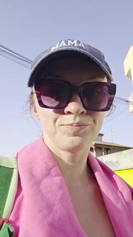 A woman in a blue cap that says "MAMA" and large purple sunglasses takes a selfie. She is wearing a white tank top and a bright pink towel is draped around her neck and shoulders. A backpack with green straps and red clasps is on her back. She appears to be outdoors, with buildings and power lines visible in the background. The lighting suggests it is daytime.