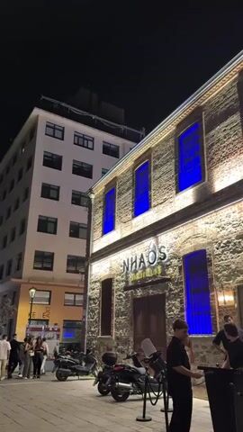 The video depicts a nighttime scene outside a building with the name "NHAOS" in large letters above the entrance. The building has a stone facade with blue-lit windows. Several people are standing around near the entrance, some interacting with each other. There are also scooters parked nearby. To the left, a taller, white building with multiple stories is visible. The street is paved and lit by streetlights.