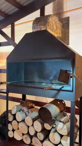 This media file showcases an outdoor barbecue setup in what appears to be a covered patio or an open-air structure. The primary object is a large, dark metal barbecue grill with a chimney-like hood. The grill is currently in use, with smoke visible inside the cooking chamber. A metal skewer with what looks like food is positioned within the grill, suggesting the barbecue is actively cooking.

Below the grill, a substantial stack of cut firewood is neatly piled, ready for use. Next to the woodpile, a metal charcoal chimney starter sits upright. The background reveals a wooden ceiling with exposed beams, and a translucent, corrugated wall that allows natural light to filter in, indicating it might be daytime. The overall impression is one of preparation for or engagement in an outdoor cooking event, likely a gathering or a meal being prepared. There is no visible text in the media. The scene suggests a rustic or traditional approach to grilling.