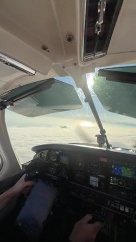 The video shows the interior of an aircraft cockpit, looking forward through the windshield. The sky is visible, with a dense layer of clouds below. Sunlight streams in from the right, creating lens flare and a rainbow effect on the windshield. In the distance, another aircraft is seen flying above the clouds. The cockpit displays various instruments and screens, and a pilot's hands are visible on the controls. The scene is one of flight above a cloud layer.