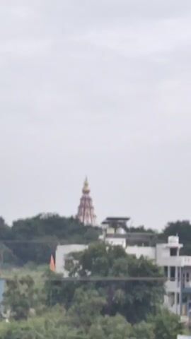 The video shows a tall, multi-tiered tower or spire, likely part of a temple, rising above a dense canopy of green trees. The sky is overcast and grey, suggesting a cloudy day. In the foreground, parts of buildings are visible, including rooftops and walls with various colors and architectural styles, indicating a residential or urban area. A lamppost with a sign is also partially visible. The camera pans slightly, revealing more of the landscape and the distant structure.