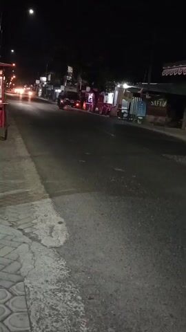 The video shows a nighttime scene of a road with several vehicles and buildings. A motorcycle is seen passing by a row of shops or stalls. Further down the road, a green truck is in motion, followed by a blue tanker truck with a large cylindrical tank. The road appears to be paved, with a sidewalk made of interlocking concrete blocks on the left side of the frame. Streetlights and lights from the buildings illuminate the scene. The overall atmosphere is that of a commercial or residential street at night.