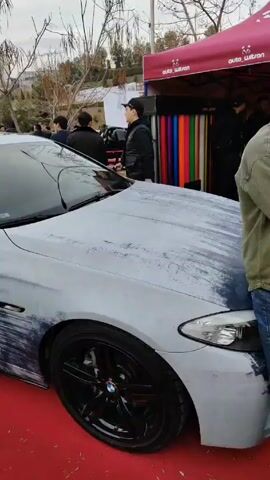 The video shows a uniquely painted BMW sedan on display at an outdoor event. The car has a white and blue, brushstroke-style paint job with red and blue stripes. The BMW is parked on a red carpet next to a display tent for "auto Watson." Several people are standing nearby, looking at the car. The weather appears to be overcast. The location seems to be an urban park or gathering space.