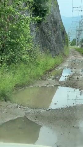 The video shows a vehicle driving on a dirt road with numerous puddles and debris. The road is flanked by a steep, rocky embankment on the left, covered in green vegetation. On the right, there are power lines and utility poles, and further in the distance, railway tracks with green train cars are visible. The sky appears overcast. The video captures the splashing of water as the vehicle navigates through the puddles.
