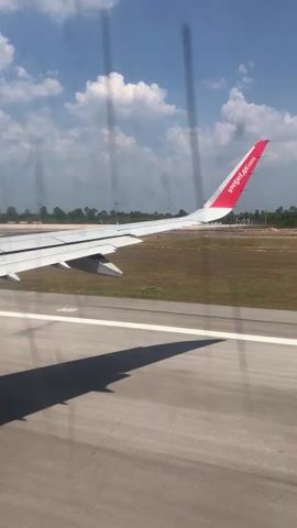 The video shows a view from inside an airplane window. A Vietjet Air plane wing is visible outside, along with a runway and grassy area. There are also some distant buildings and trees, and a blue sky with scattered clouds. The plane appears to be moving along the runway.