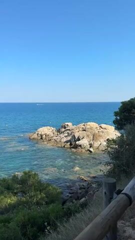 The video shows a scenic coastal path with a wooden railing alongside. To the left, the turquoise sea is visible with some rocks jutting out of the water. Lush green trees and bushes line the path and the coastline. The sky is a clear, bright blue. The path itself is made of dirt and gravel, and it curves gently as it ascends. The camera is moving along the path, offering a dynamic view of the landscape. The overall impression is one of a peaceful and beautiful natural environment.