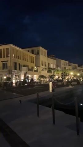 This media file captures a nighttime scene in Kumbor, Montenegro. The setting is a waterfront promenade lined with modern, brightly lit buildings that appear to be hotels or upscale residences, possibly with shops or restaurants on the ground floor. The architecture is contemporary, featuring balconies and large windows.

Several people are visible on the promenade, some walking and others sitting on benches. Their activities suggest a relaxed evening atmosphere, perhaps enjoying a stroll or dining out. The lighting from the buildings and streetlamps illuminates the paved walkway, creating reflections on the wet surface, suggesting recent rain or a general dampness in the air.

The sky is dark, indicating nighttime, with some clouds visible. In the background, the silhouette of mountains can be seen against the night sky, a common geographical feature of the region. The overall mood is tranquil and inviting, a typical evening ambiance for a coastal resort town. No visible text is present in the media file.