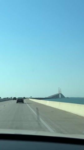 The video shows a vehicle driving on a highway, approaching a large bridge. The sky is clear and blue. To the right of the road is a body of water, possibly an ocean or bay.  The bridge is visible in the distance. As the car continues to drive, the bridge disappears from view and the video focuses on the water and the sky.