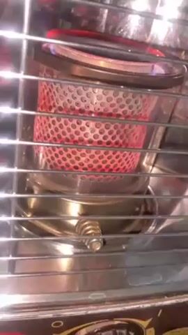 The video shows a close-up of a kerosene heater in operation. The heater has a circular, perforated metal mesh that glows bright red with heat. Blue flames are visible at the top edge of the mesh. The heater is surrounded by a chrome cage with horizontal bars, likely for safety. A brass knob is visible on the front of the heater. The camera is still and stable, focusing on the illuminated core of the heater.