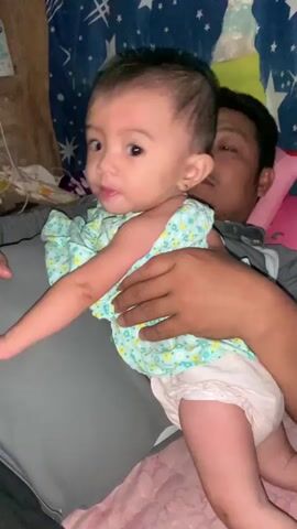 A man holds a baby in his arms. The baby is wearing a light blue and green floral print onesie and a diaper. The man is wearing a gray shirt. The background appears to be a bedroom with a blue and white star-patterned blanket or curtain. The lighting is dim. The baby is looking towards the camera and sticking out its tongue. The man is looking towards the baby.