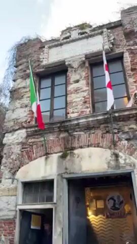 The video shows the exterior of a weathered brick building. The building has two visible windows with dark frames. Above the windows, there is a stone plaque with Latin text engraved on it, framed by decorative stonework. The building's facade shows signs of age and wear. An Italian flag is on the left and a white flag with a red cross is on the right, both positioned between the ground and the windows. A doorway leads into the building, and a person is visible inside. There are also people near the doorway outside, moving around. The sky is blue with some white clouds. The camera pans up from the doorway to show the entire facade and flags.