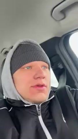 Here's a description of the video:

A young man with light eyes is sitting in the driver's seat of a car. He is wearing a grey hooded sweatshirt under a black jacket with white stripes on the sleeves, and a grey beanie. He is wearing a seatbelt. He is looking straight ahead as if he is driving. Music is playing. The view out of the car window shows an overcast sky.