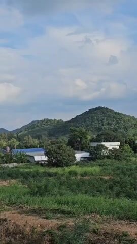 This is a vertical video showing a landscape with hills and vegetation under a cloudy sky. In the foreground, there is dry ground with some grass. Behind it, there is a field of green plants. In the middle ground, there are more green plants and trees, with some buildings visible, including structures with white roofs and one with a red roof. In the background, there are several lush green hills, the tallest one being in the center. A tall antenna structure is visible on the right side of the largest hill. The sky is blue with scattered white clouds. The video pans slightly to the right.